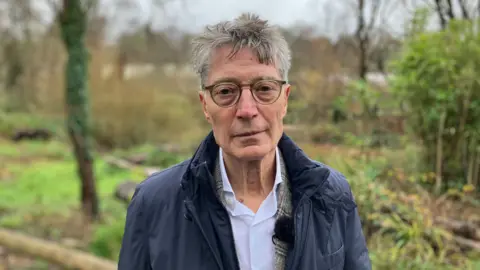 Kirk England/BBC Dirk Lips has a blue jacket and an open necked white shirt. He is wearing glasses and is looking directly at the camera.