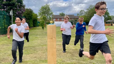 Five boys in grey trousers and white polo shirts are running in a school field. One is wearing a blue jumper. They are running past a wooden post with a football figurine on top of it.
