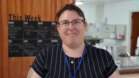 Shaun Whitmore/BBC Louise Daglish standing in front of a serving hatch in a care home. She has short dark brown hair, is wearing glasses and a blue T-shirt with fine white stripes. Behind her is a blackboard menu. Through the hatch is a woman in chef's whites. 