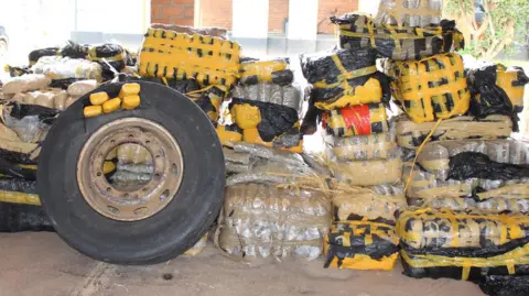 A large pile of seized packages, tightly wrapped in black and clear plastic and secured with bright yellow tape, with smaller yellow-wrapped blocks stacked on top. A large vehicle tire leans against the front of the pile.
