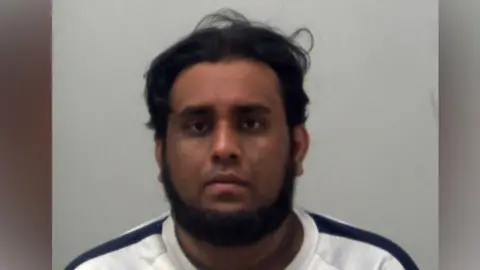 Oaveed Rahman is wearing a white top with a black stripe along both shoulders. He has a beard and he is looking at the camera. It is a custody photo.