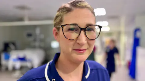 Nikki Fox/BBC Senior sister Lauren Jakes stands in the critical care department at the Norfolk and Norwich University Hospital  The background is blurry but you can see a bed to the left of her and another nurse to the right. She is wearing a blue uniform with her hair plaited back and tortoiseshell glasses.