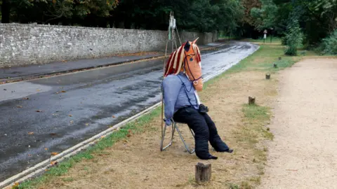 A scarecrow with a horse's head on a stool. The stool is on a grass verge at the side of a road.  A long stone wall and trees can be seen in the background. 