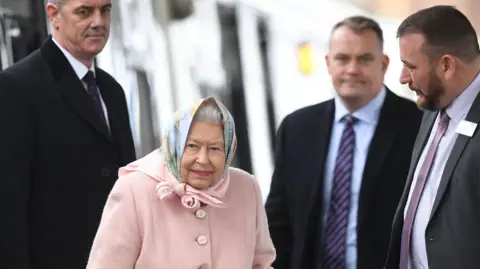 PA Queen Elizabeth II stands on a station platform surrounded by three men in suits. She wears a pale peach coat with matching peach and pale blue headscarf.