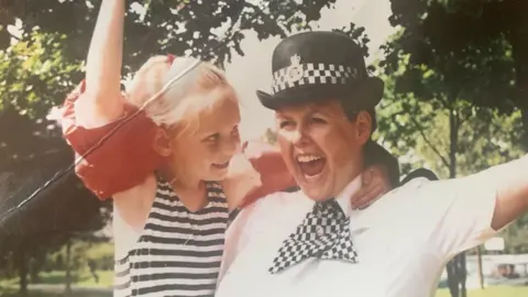 A woman in a police officer's uniform and hat grins with her arm in the air, while hugging a young blonde girl in red armbands who smiles at her.