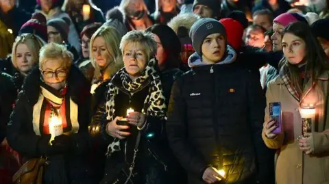 Crowds of people at a vigil in Banbridge. Some at the front are holding candles. At the front are three women and a young man. Two of the women have blonde hair and are wearing colourful scarves. The young man is wearing a dark jacket and a dark hat. The other woman has dark hair and a light coat.