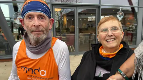 Anthony Cox wearing an orange high vis MND vest and a white top underneath it. He also has an orange MND head band on and has light facial hair. He is next to his wife Tina who is smiling as she looks slightly to the right of camera. 