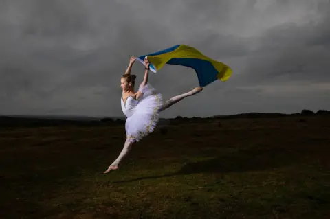 Joann Randles Accompanied with the Ukrainian flag, award-winning, young ballerina, Kateryna Andrushyna, 12, proudly demonstrates her skills for Ukraine Independence Day. Kateryna and her family are refugees from Boryspil, a city near Kyiv, that was bombed on the first day of the Russian Invasion,