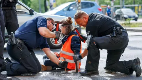 HANNIBAL HANSCHKE/EPA-EFE/REX/Shutterstock Police officers work to free a Letzte Generation (Last Generation) climate activist after he glued himself to the asphalt during a climate protest in Berlin, Germany, 22 May 2023
