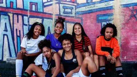 Gustavo Oliveira/WBR Photo Six girls pose for the camera against the graffiti-decorated wall of their training pitch