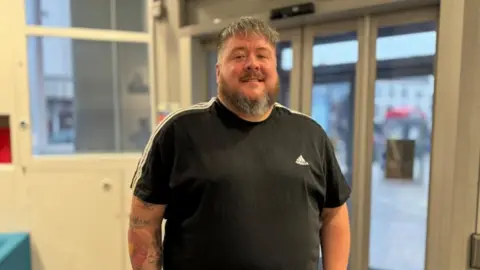 Rob Constantin/BBC Man wearing a black top with a brown beard standing by a door - some tattoos are visible on his arms