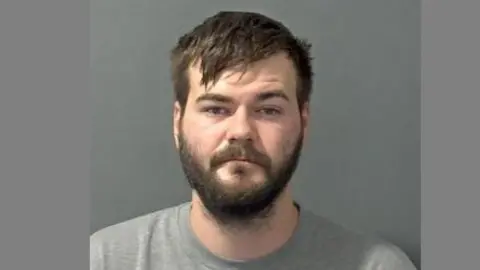 Bedfordshire Police Police "mug shot" of a man with a dark beard wearing a grey t-shirt