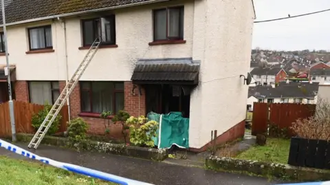 Pacemaker A cream and red brick semi-detached house which sits on a hill, with loads of houses behind. Police tape is visible at the forefront of the picture. There is a green cover over the front door, the windows look black and there is a ladder going up into a first floor window.