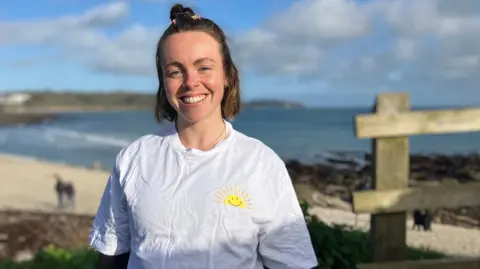 A woman smiles on a beach in Cornwall wearing a white T shirt with a golden sun on it 