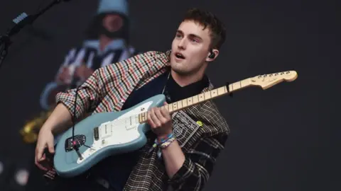 Sam Fender in a chequered open shirt with light blue guitar on stage.