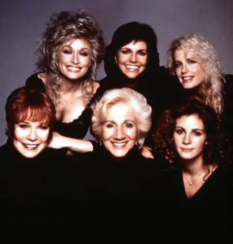 MOVIESTORE COLLECTION LTD via Alamy Shirley Maclaine, Dolly Parton, Olympia Dukakis, Sally Field, Julia Roberts and Daryl Hannah smile in a group photo, They all wear black. 