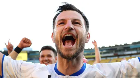 Tommy Smith celebrating on the pitch after an Auckland City game. He has a short beard and brown hair and is wearing a white top with a blue collar.