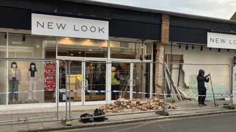 Vikki Irwin/BBC A New Look shop that has been cordoned off which police tape and fencing after a collision. One person can be seen with a brush sweeping up the front of the building. 