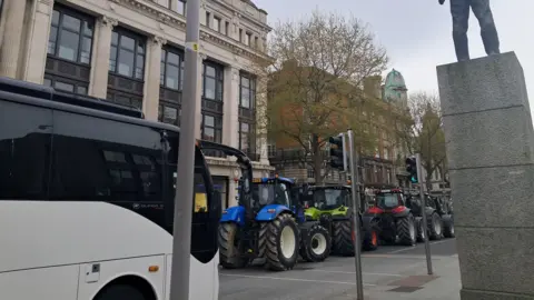 Several tractors parked on a city street. Behind them is a white bus. On the right is the bottom of a statue.