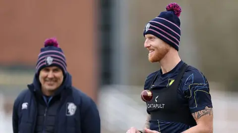 Ben Stokes in T-shirt and woolly hat, throws a red cricket ball up as he prepares to bowl in training as coach Ryan Campbell, in navy jacket and woolly hat watches on from a slight distance