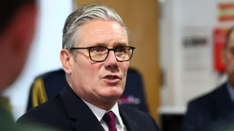 Keir Starmer, who wears his short grey hair combed to the side and black-rimmed glasses, is seen speaking to trainee pilots at an RAF base