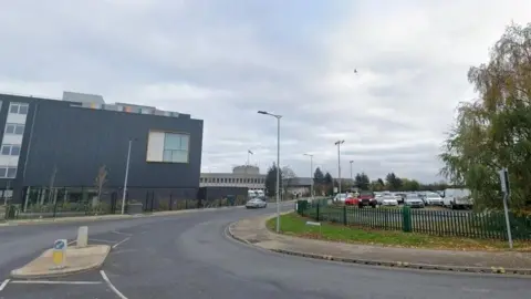 Google streetview image of a road, just coming off a junction, and curving round to the right. There is a car park on the right hand side and a large dark grey building on the left of the road. A brick building on the left is the police station.