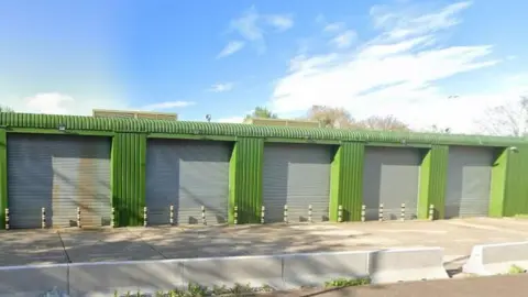 The exterior of the former recycling centre. The  building is mainly green with grey shutters and bollards in front of the building.  