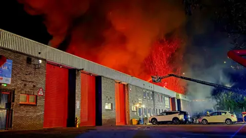 South Yorkshire Fire and Rescue A large warehouse building on fire with three cars parked outside. There are huge red flames coming from the top of the building while firefighters on a cherry picker shoot water down