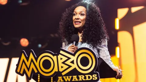 Getty Images Kanya King speaking on stage at a lectern with a large yellow Mobo Awards logo at the 2025 ceremony
