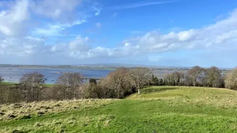 A view of across the Somerset Levels on a clear sunny day. Green fields and slightly bare trees can be see in front of a flooded plain.