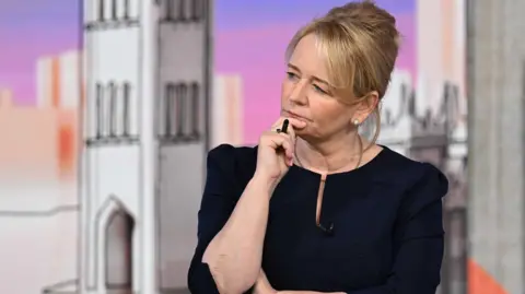 Unite General Secretary Sharon Graham has blonde hair tied back and is wearing a navy top. She has her hand on her chin and is holding a pen and looking away from the camera. She is also wearing a small microphone.