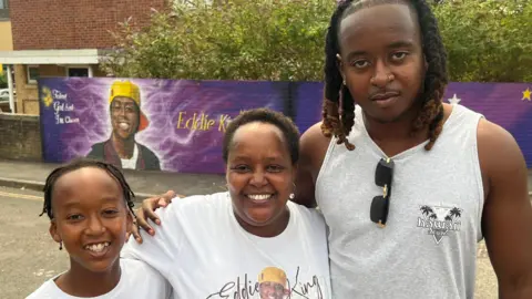 Irene stands between her son Elliot and a younger child. She and the younger child are wearing white t-shirts with a picture of Eddie on them and his name. The three are holding each other and smiling brightly in front of the mural. 