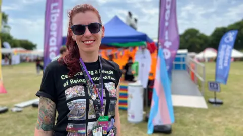 Kaye stands outdoors at an event, wearing a black t-shirt that reads 'Celebrating a Decade of Being Proud of You' with images printed on it. She has two lanyards and an ID badge. Behind them are two tall purple 'Liberate' flags, a blue tent, and colourful decorations including a rainbow flag and a transgender pride flag. The scene is set on a grassy field with other tents and people in the background under a partly cloudy sky.