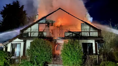 A large fire at a house. There is a orange blaze coming from the building and a lot of smoke. 