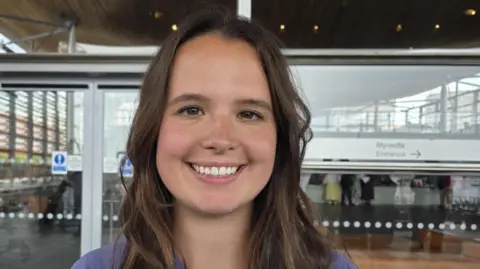 BBC Zaynub Akbar smiling outside the Senedd in Cardiff Bay with glass entrance door and windows behind her. She has long brown hair and smiles and wears a mauve shirt
