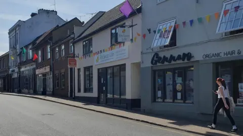 Gennaro Borrelli A street in Leighton Buzzard, showing rows of shops, and a road, with a woman walking along it. There is bunting on the buildings. There are no cars on the road.