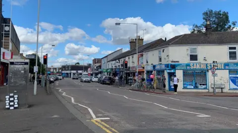 Shariqua Ahmed/BBC A road is in the middle with buildings either side. On the left is brown bricked building with red painted windows on the bottom. On the right is a white building which has a road leading down beside it.