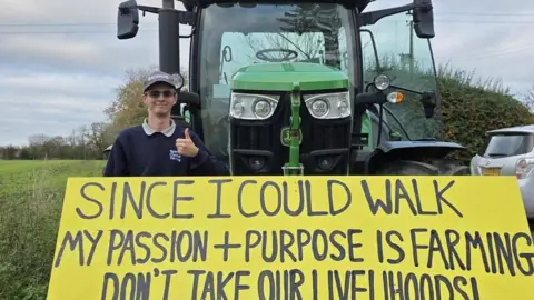 Supplied A young man wearing a cap and a navy jumper standing next to a tractor with a large yellow sign. He is smiling and has his thumb up. 
