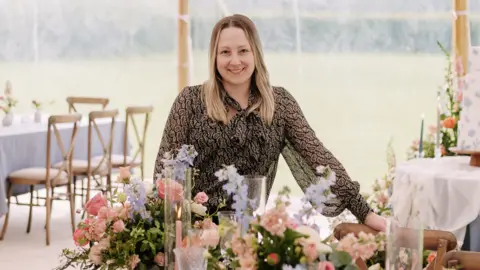Stu Tucker A woman stood in a marquee, behind some florals. She is looking into the camera smiling.