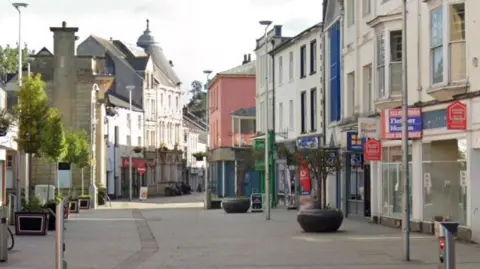 Google A section of George Street in Pontypool shows shops including Greggs on the right hand side, with street furniture including large plant displays.