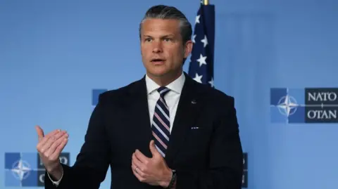 Hegseth gestures with both of his hands as he stands in front of a Nato branded blue backdrop at a summit. He is wearing a dark blue suit, white shirt and white, blue and pink striped tie.