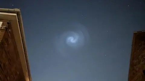 A spiral light set against a dark night sky framed by the eaves of two bright houses.