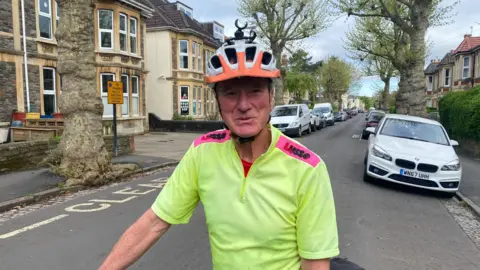 Tacey Bain Photo of David in a fluorescent t-shirt and cyclist helmet standing in the middle of a quiet road. There are buildings with bay windows on the street and parked cars behind David. 