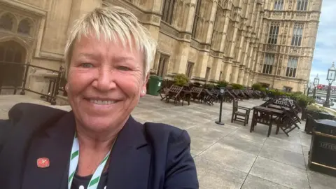 Rachel Taylor A woman in a selfie in front of the Houses of Parliament. She has short blonde hair, and she is wearing a dark  blue suit jacket.