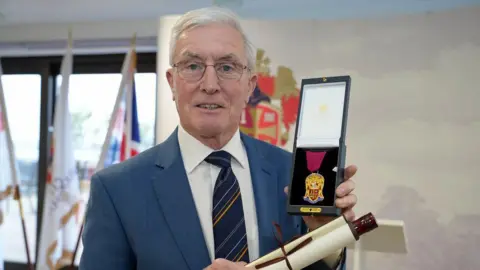 An older man in a blue suit and white shirt, holding a scroll and a small gold medallion. He stands in a room with white patterned walls with a crest painted on them and tall windows, with flags on poles in front.