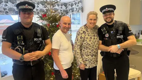 Two police officers in black uniform standing with a couple inside a home with a Christmas tree in the back. A man is wearing a white t-shirt. He is bald. The woman is wearing a leopard print fleece. They appear to be standing inside a kitchen