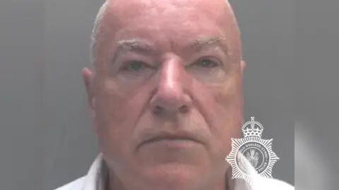 A mugshot of a man with shaven white hair, looking directly into the camera with a neutral expression. 