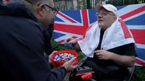 A bishop in black religious clothing holds an open red tub of Celebrations chocolates down to a sitting protester, a man with a white cap who has a St George's flag around his shoulders and a Union Jack banner pinned to the fence behind him.