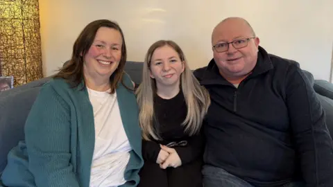 Isabella Verona/ BBC Ava and her parents sit together on a sofa in a living room. The woman on the left is wearing a textured, teal-coloured cardigan over a light top. Ava sits in the middle and has long, light-toned hair and is dressed in a dark outfit, with her hands resting in their lap. The man on the right is wearing a dark zip-up top. Behind them, a tall woven-style floor lamp glows warmly, and framed pictures are visible on a side table. A wall with light-coloured paint and a decorative picture hanging above the group forms the background.