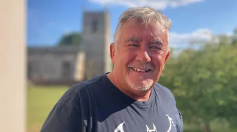 Gary Cawkwell smiles into the camera. He has short grey hair, a grey goatee beard and is wearing a black t-shirt with a Batman logo. Gary is standing in the sunshine and there is a church building in the background and a large green tree on the right of the image.